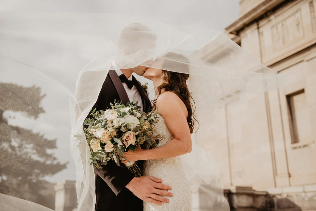 Romantic wedding couple kissing under a flowing veil, bride holding a soft blush and white bouquet, with warm sunlight and elegant architecture in the background