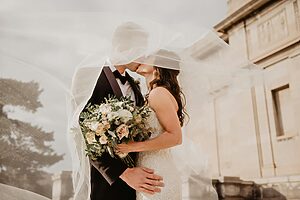 Romantic wedding couple kissing under a flowing veil, bride holding a soft blush and white bouquet, with warm sunlight and elegant architecture in the background