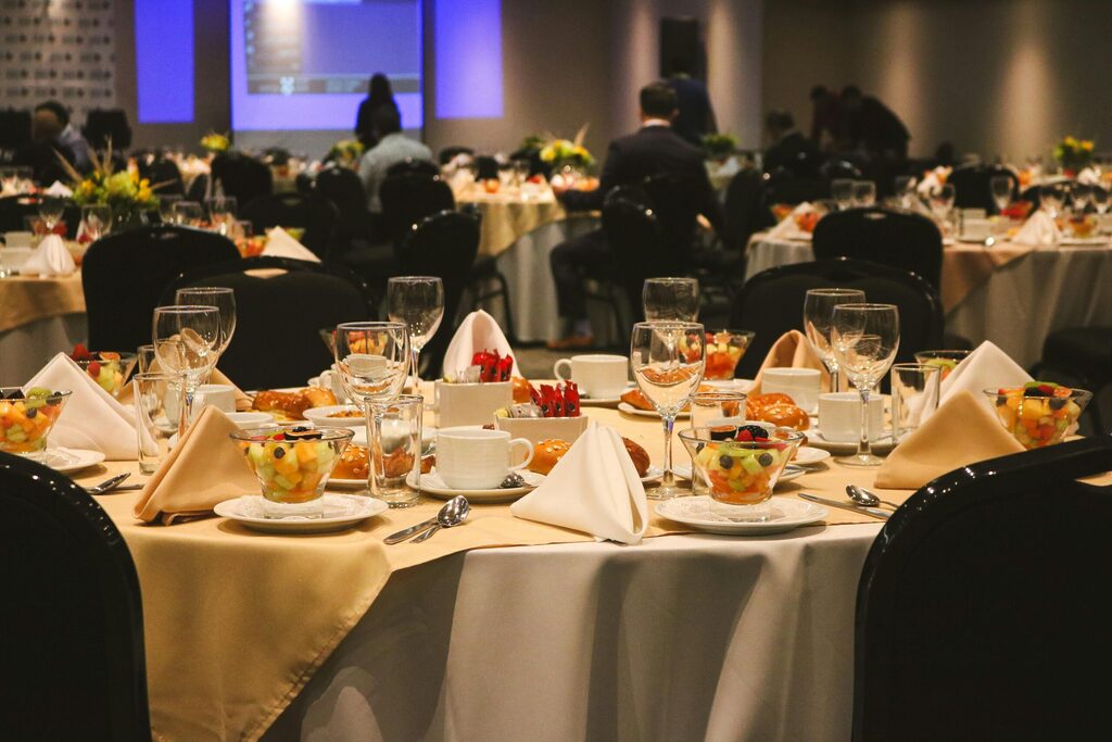 Formal event banquet setup with round tables, white tablecloths, neatly folded napkins, glassware, fruit starters and bread rolls, with guests seated in a conference-style dining hall.