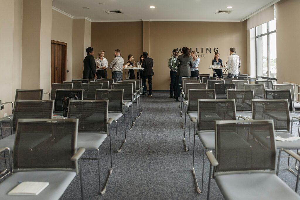 Conference room with rows of empty chairs facing a presentation area, while attendees network and chat around high tables at the back of a modern hotel meeting space.