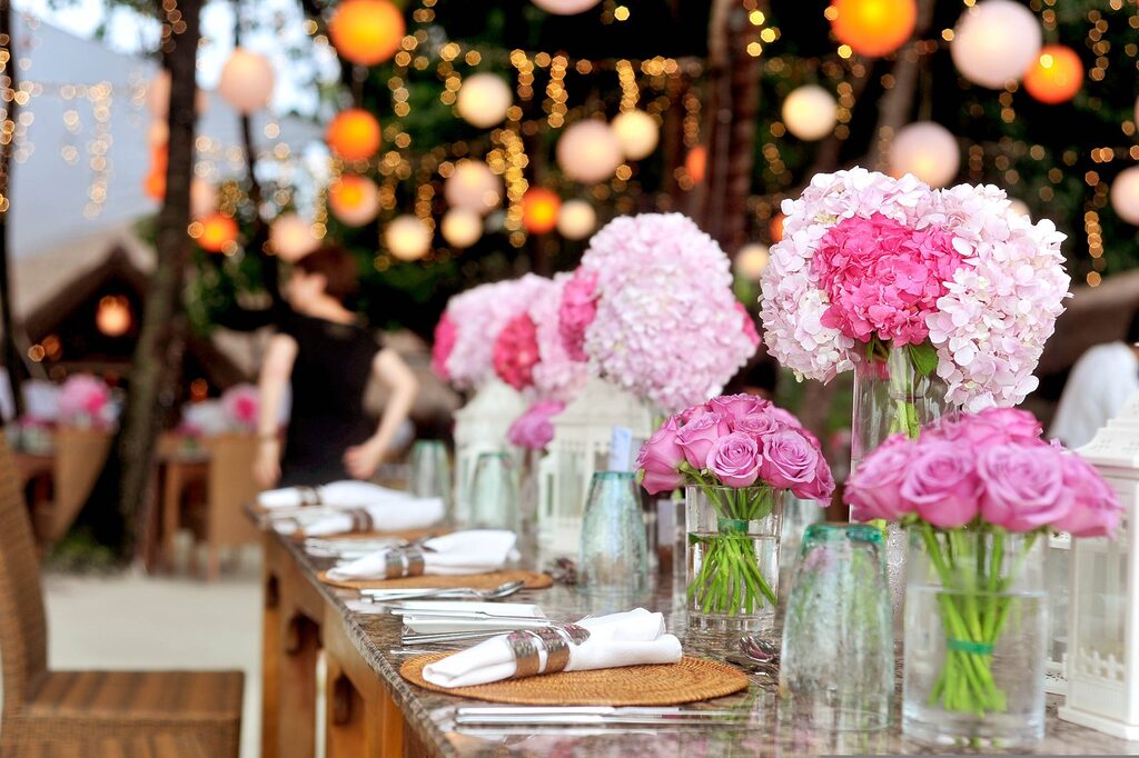 Elegant outdoor dining table decorated with pink rose and hydrangea centrepieces, set with glassware and napkins, under warm hanging lights and lanterns in a garden setting.