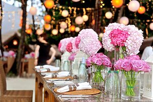 Elegant outdoor dining table decorated with pink rose and hydrangea centrepieces, set with glassware and napkins, under warm hanging lights and lanterns in a garden setting.