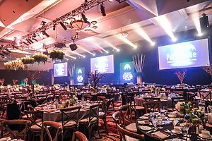 Large corporate event venue with banquet tables, stage lighting, LED screens and formal dining setup for conference or gala event
