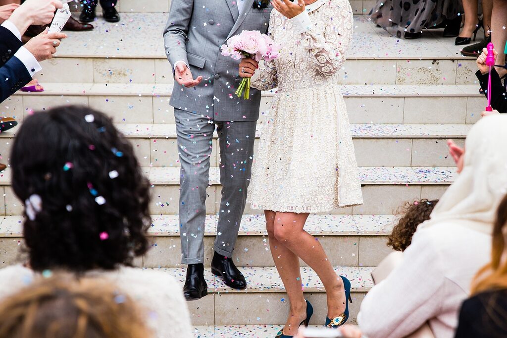 Bride and groom walking down stone steps after their wedding ceremony as guests throw colourful confetti, bride holding a bouquet of pink flowers.