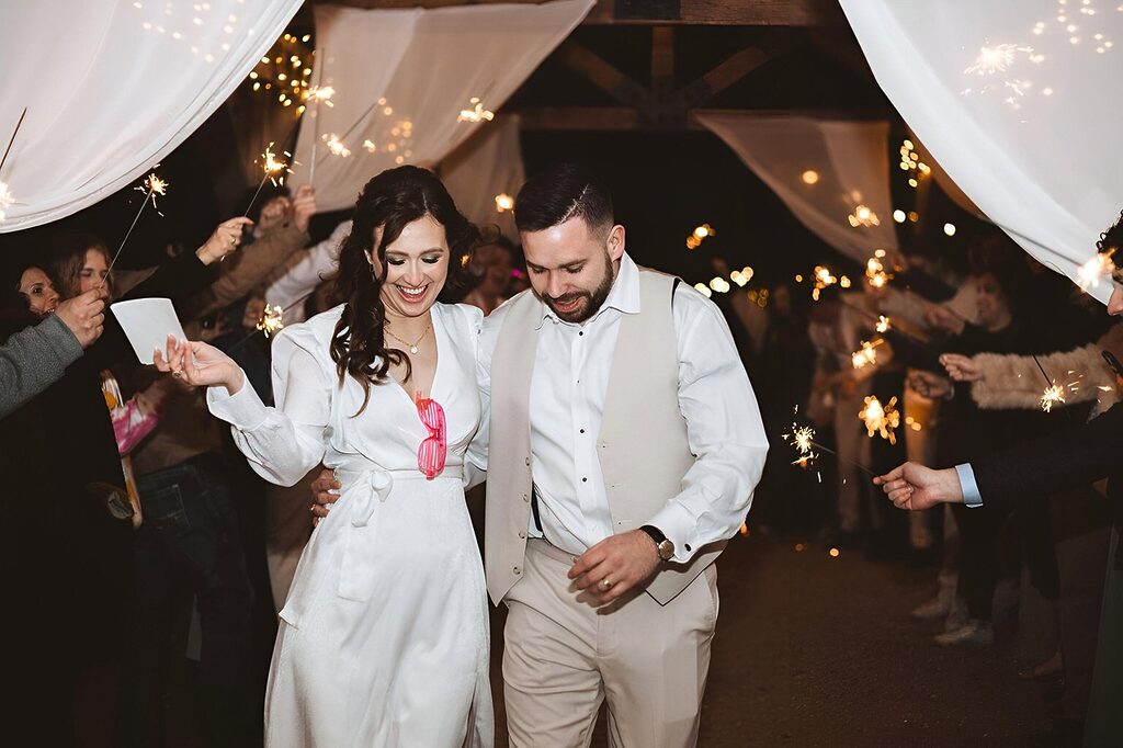 Bride and groom walking hand in hand through a sparkler send-off at night, surrounded by guests celebrating under white draped canopy.