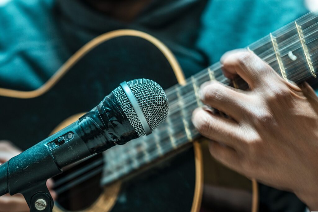 Close-up of a musician playing an acoustic guitar while singing into a microphone, focusing on hand placement on the fretboard and studio recording setup
