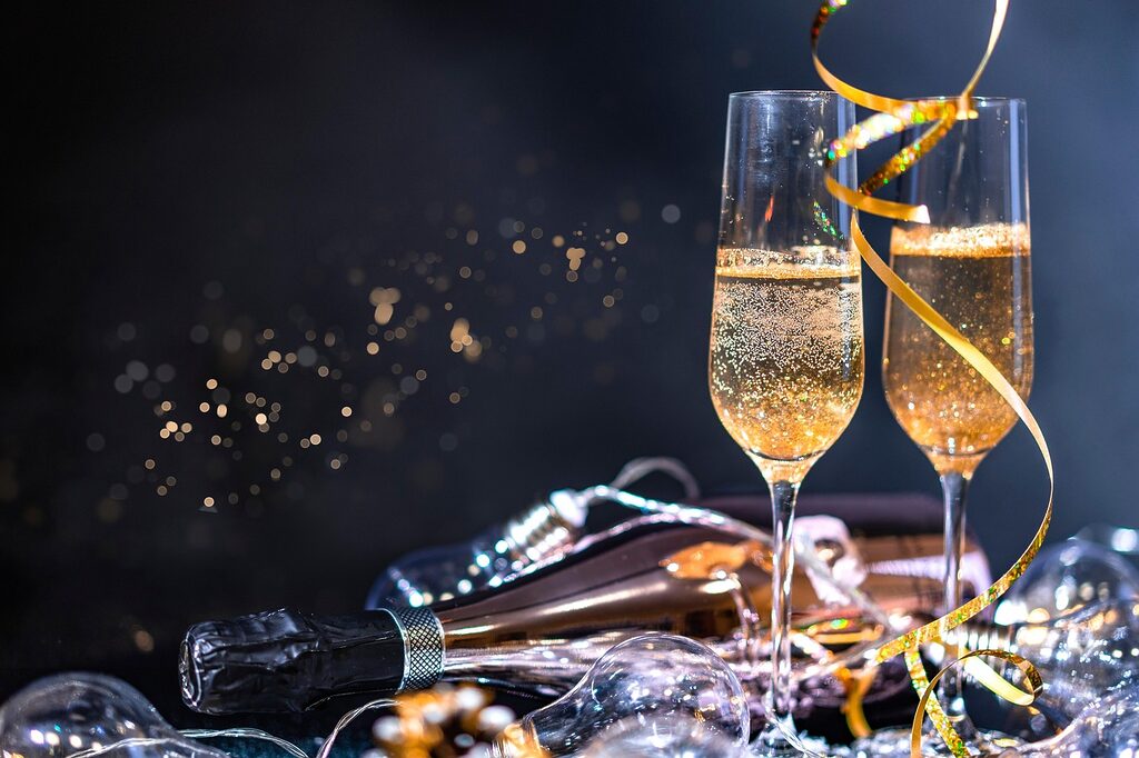 Two champagne flutes with sparkling wine beside a bottle, party lights and gold ribbon on a festive table.