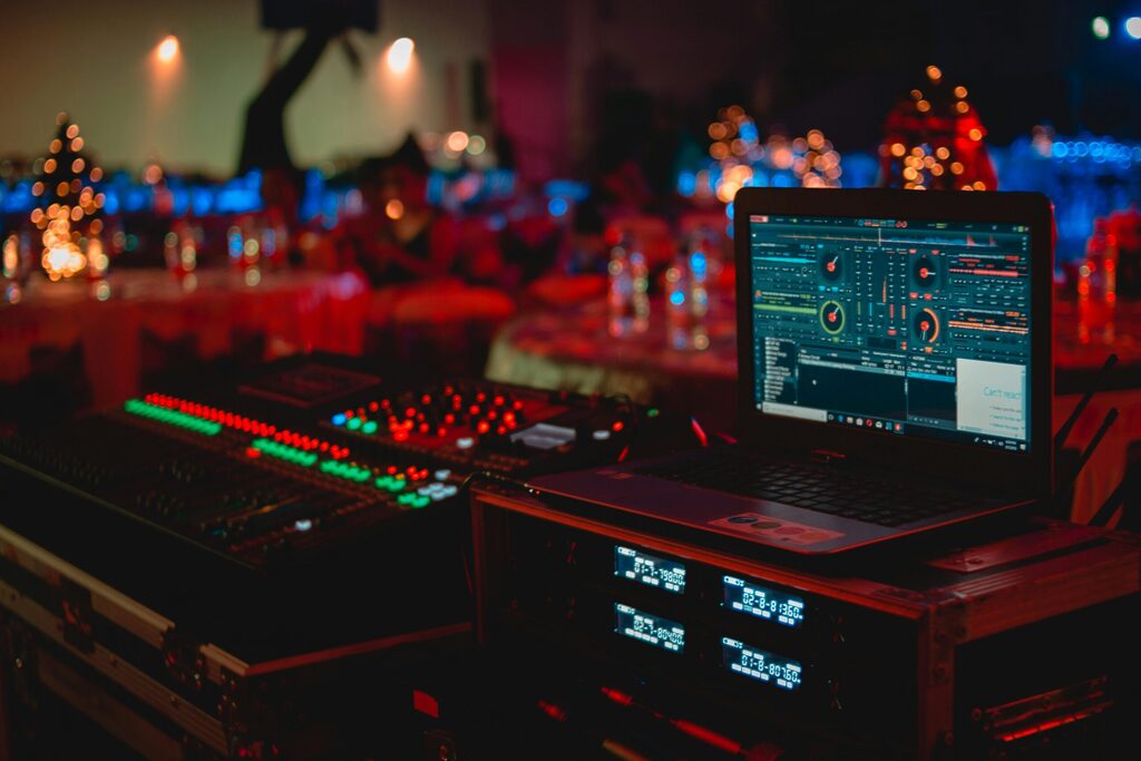 Professional DJ setup at an event, featuring a laptop running mixing software, audio mixer with illuminated controls, and wireless receivers, with softly lit banquet tables and decorative lights blurred in the background.