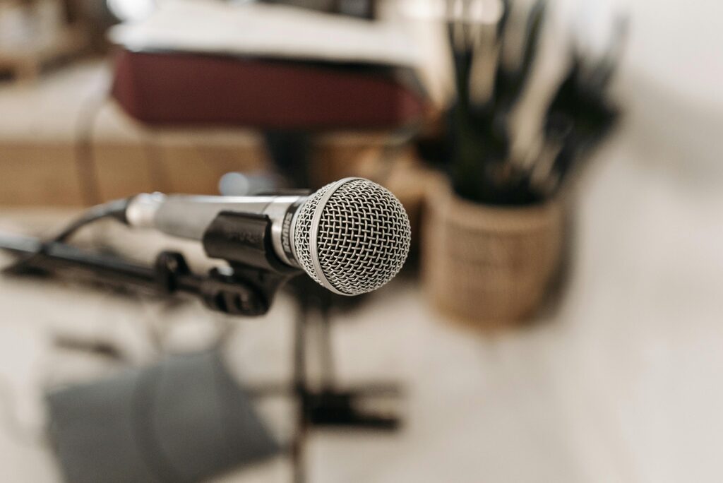 Close-up of a microphone on a stand in a meeting or presentation setting, with a blurred background showing a table and indoor decor.