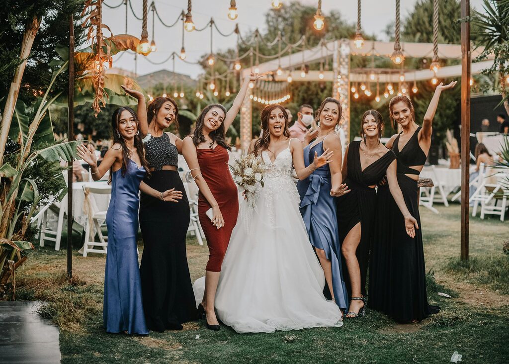 Bride celebrating with bridesmaids in elegant dresses at an outdoor wedding reception, with string lights, greenery, and a festive garden setting in the background