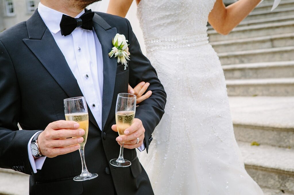 Bride and groom celebrating on stone steps, groom in black tuxedo holding two champagne glasses beside bride in beaded white wedding dress.